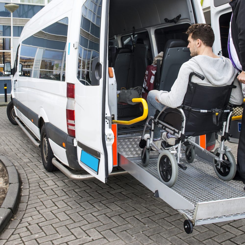 The driver of a wheel chair taxi, helping a disabled man in a wheel chair, using the lift in the back of his mini van